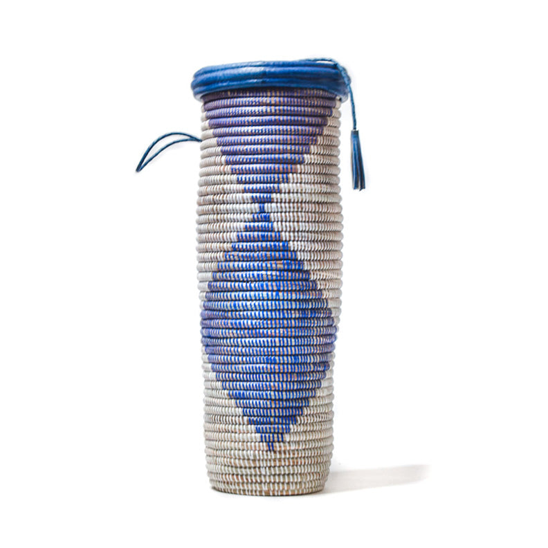 Jummy Basket on white. Tall, skinny, rounded woven basket with large blue diamond pattern. And a blue leather trimmed, tasseled lid.